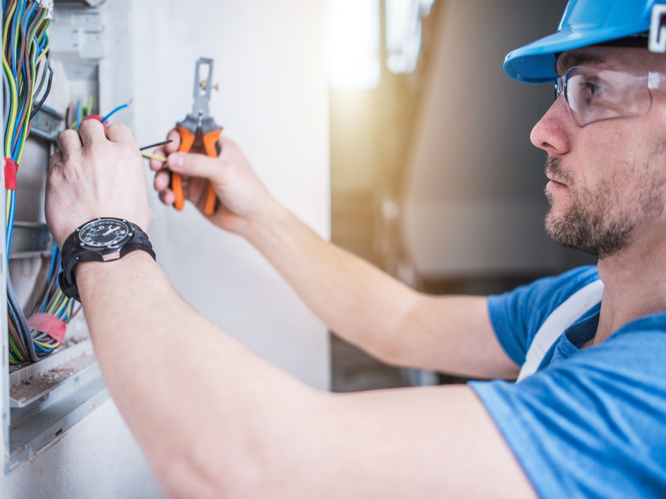 An electrician in a hard hat and safety glasses wiring a wall box, pliers in hand