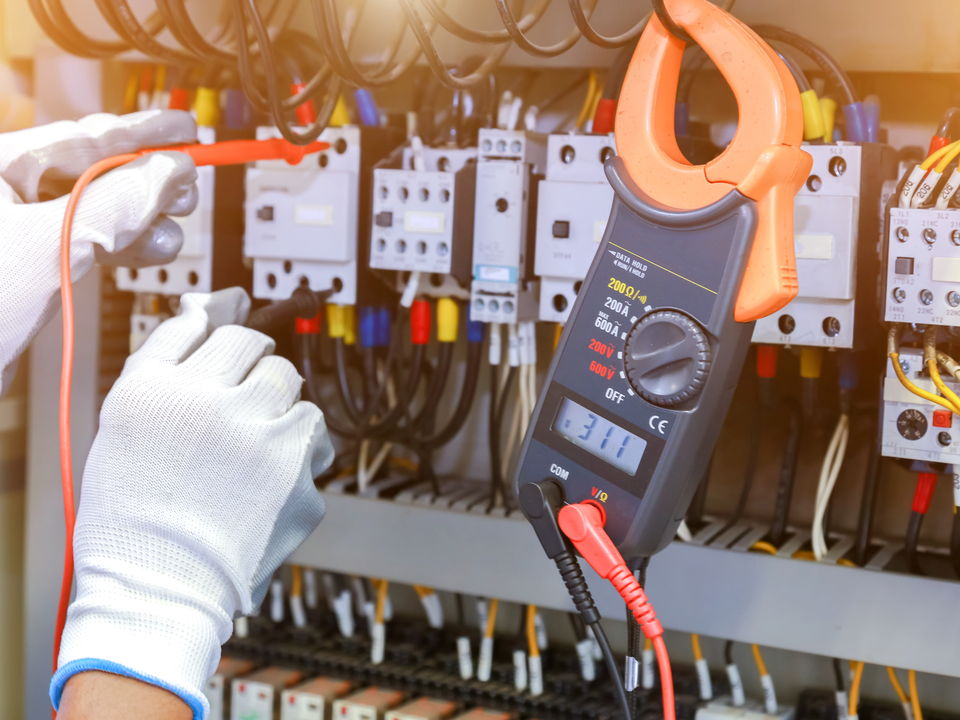 A gloved hand holding a clamp meter against the wiring of a switchboard