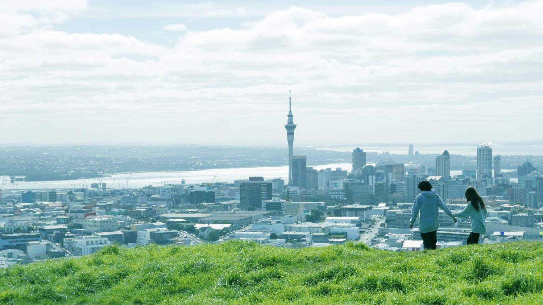 The Auckland skyline and Sky Tower from the grassy slope of Mount Eden, a couple walking in the foreground