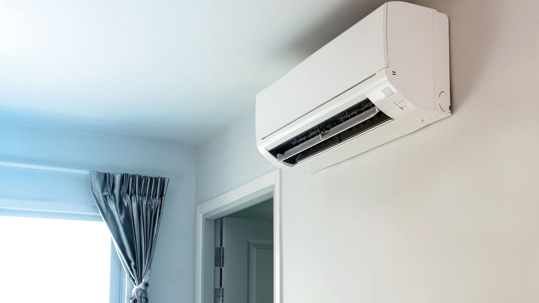 A white wall-mounted heat pump indoor unit fitted high on a wall beside a doorway, a curtained window to the left