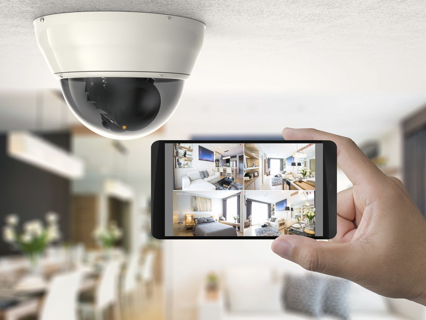 A dome security camera on a ceiling, with a hand holding a phone that shows the live camera view of the room