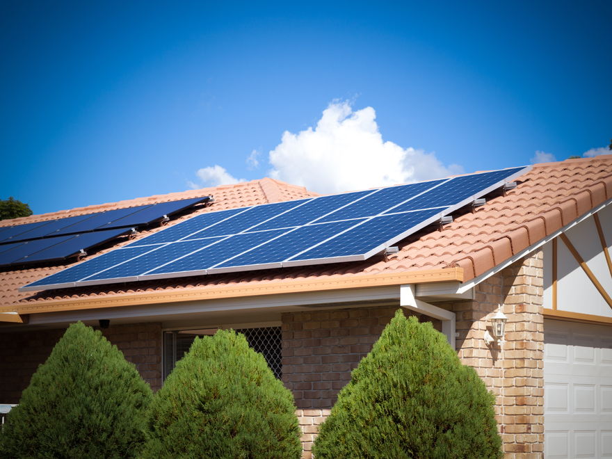 Solar panels on the tiled roof of a single-storey brick house, conifers in front